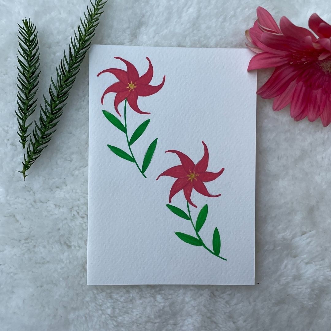 A hand painted greeting card with various flower stalks painted on a white card placed on a a fur cloth with decorative flowers in the background 