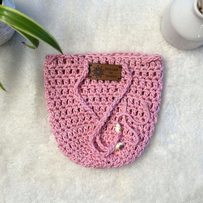 Pink hand crocheted pot cozy with a drawstring and a faux leather olive leaf label placed on white fur fabric 