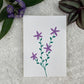 A hand painted greeting card with various flower stalks painted on a white card placed on a a fur cloth with decorative flowers in the background 