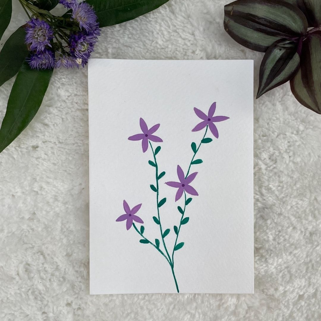 A hand painted greeting card with various flower stalks painted on a white card placed on a a fur cloth with decorative flowers in the background 
