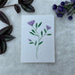 A hand painted greeting card with purple lotus flower stalks painted on a white card placed on a a fur cloth with decorative flowers in the background 
