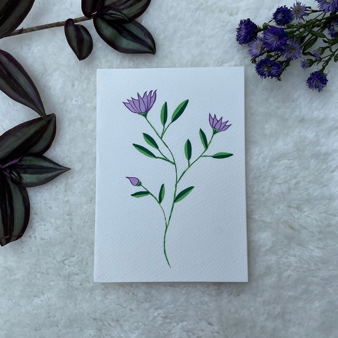 A hand painted greeting card with purple lotus flower stalks painted on a white card placed on a a fur cloth with decorative flowers in the background 