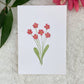 A hand-painted greeting card with a tiny pink floral gouache illustrations on a white background, displayed on a fluffy surface with a small branch of leaves to the side.