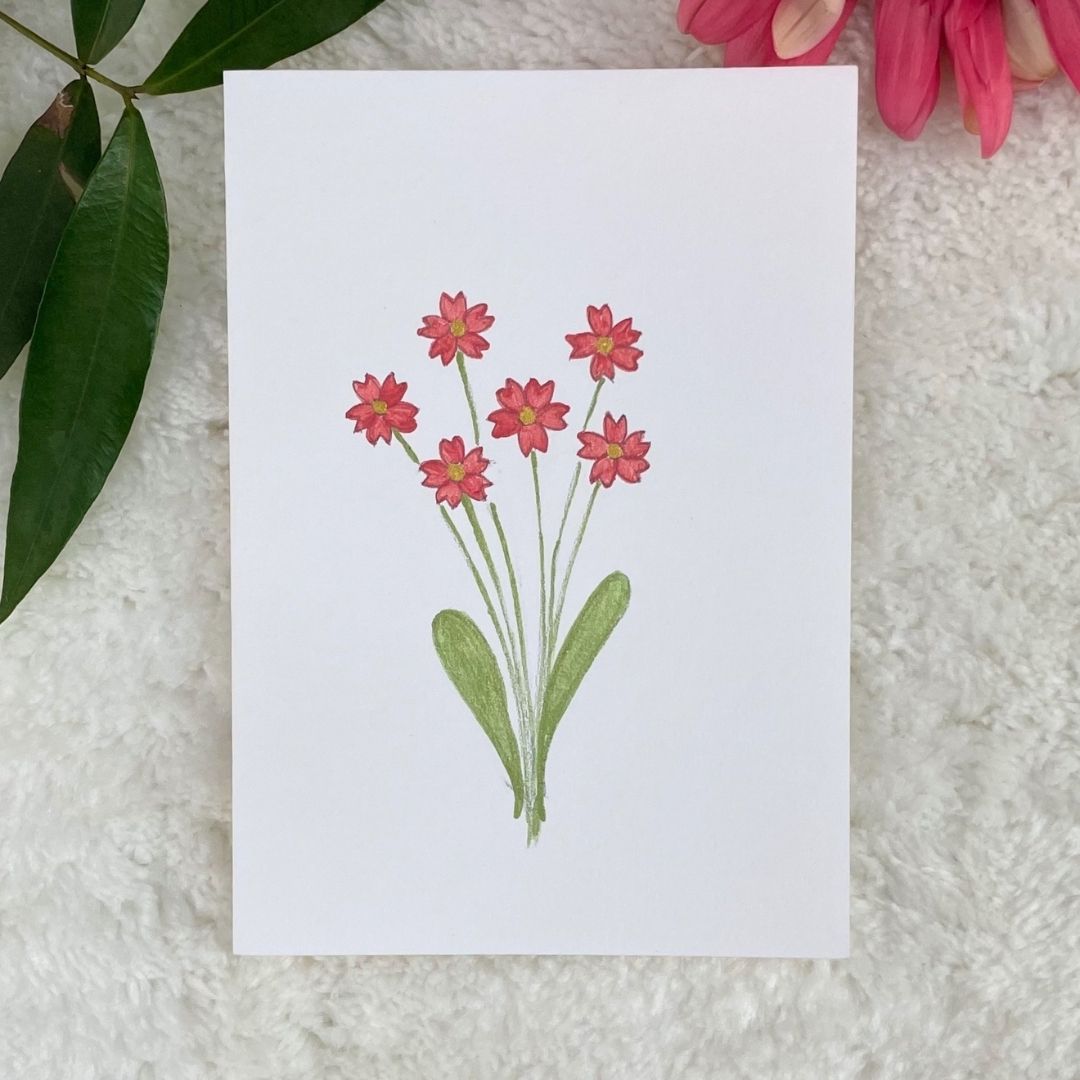 A hand-painted greeting card with a tiny pink floral gouache illustrations on a white background, displayed on a fluffy surface with a small branch of leaves to the side.