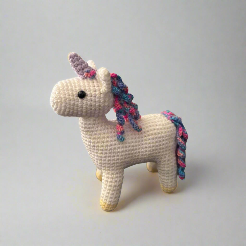 Crocheted white unicorn plushie with a purple horn and rainbow coloured mane and tail standing on a white surface 