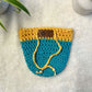 Yellow and blue hand crocheted pot cozy with a drawstring and a faux leather olive leaf label placed on white fur fabric 