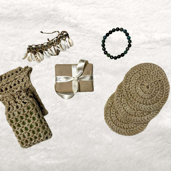 Bath set comprising of 2 soap avers one with a soap inside it, 3 face scrubies placed on top of each other, a soap wrapped in brown paper and tied with a ribbon and trinkets in the background 