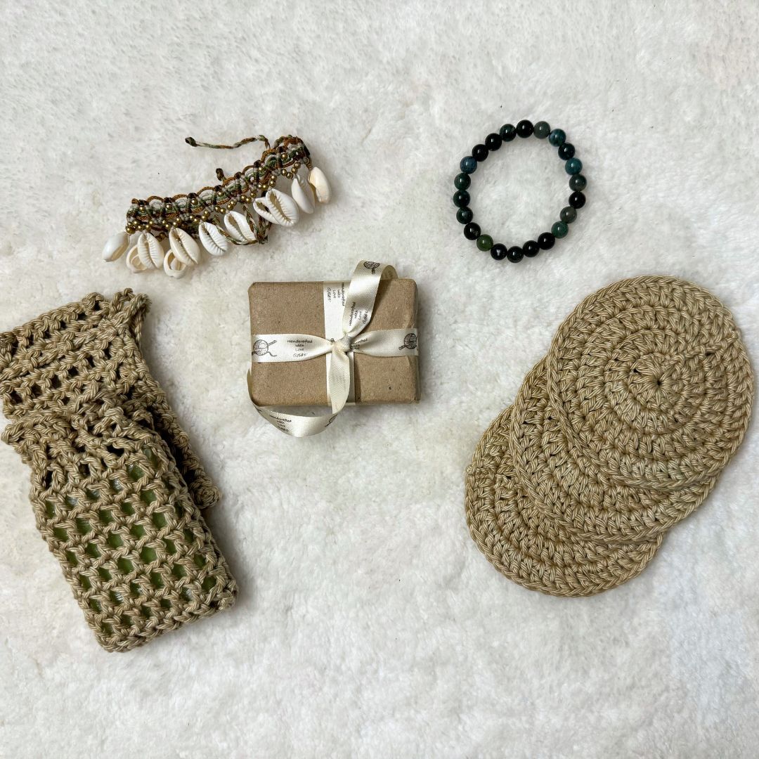 Bath set comprising of 2 soap avers one with a soap inside it, 3 face scrubies placed on top of each other, a soap wrapped in brown paper and tied with a ribbon and trinkets in the background 