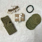 Bath set comprising of 2 soap avers one with a soap inside it, 3 face scrubies placed on top of each other, a soap wrapped in brown paper and tied with a ribbon and trinkets in the background 
