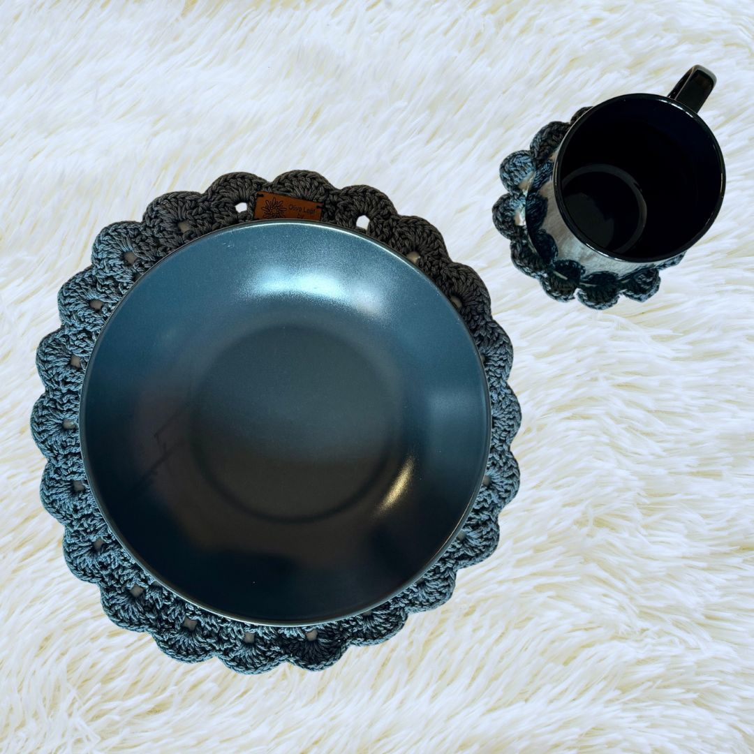 Grey hand crocheted place mat with a gey plate on it and a coaster with a cup on it placed on a soft white cloth 