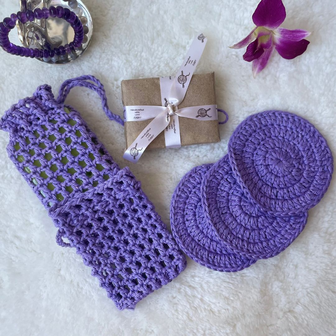 Bath set comprising of 2 soap avers one with a soap inside it, 3 face scrubbies placed on top of each other, a soap wrapped in brown paper and tied with a ribbon and trinkets in the background 