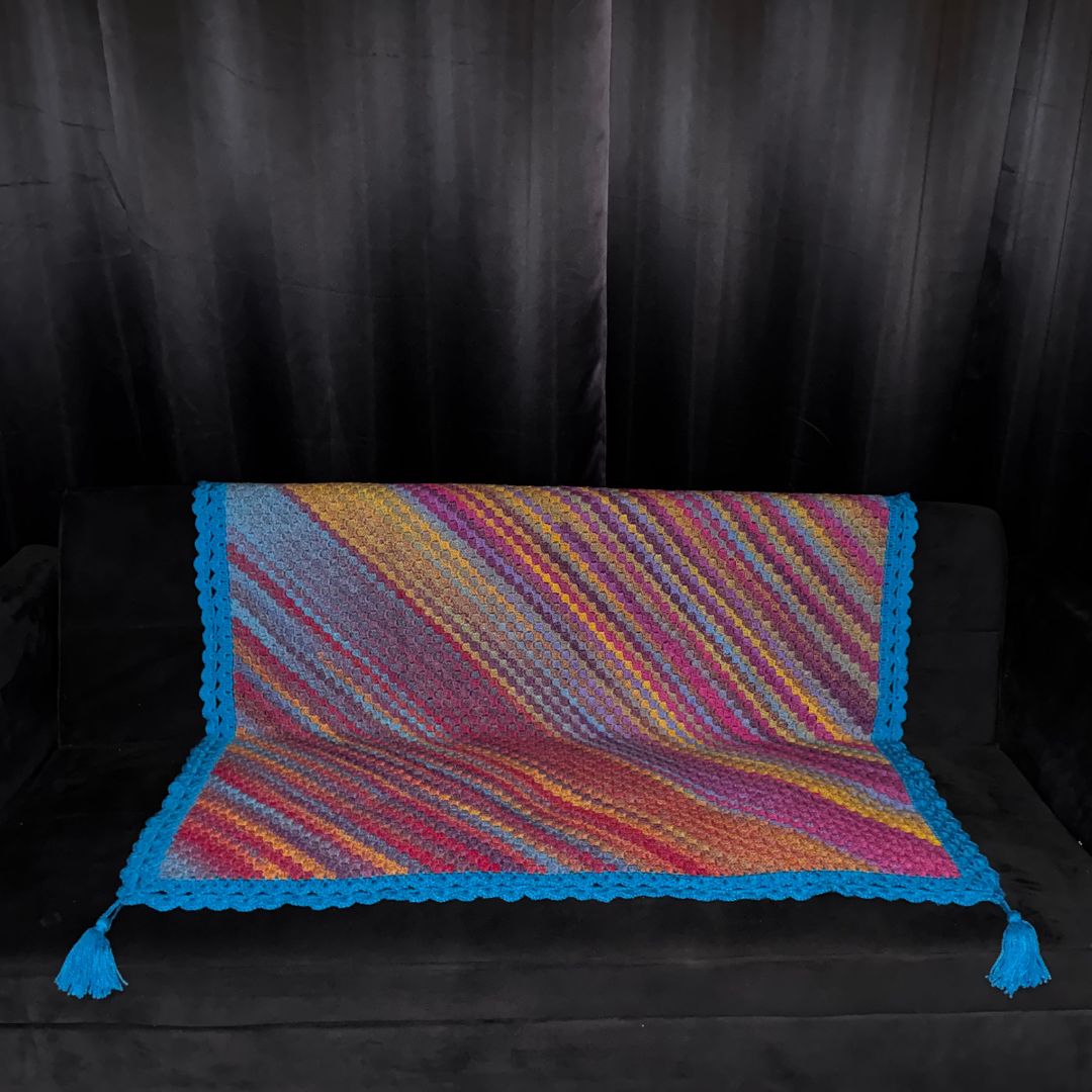 Colorful crocheted sofa throw with a bright blue border and blue tassels placed on a black sofa with black curtains in the background 