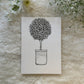 Plant with tiny leaves along with a pot sketched in black and white on a greeting card placed on a soft white cloth with flowers in the background 