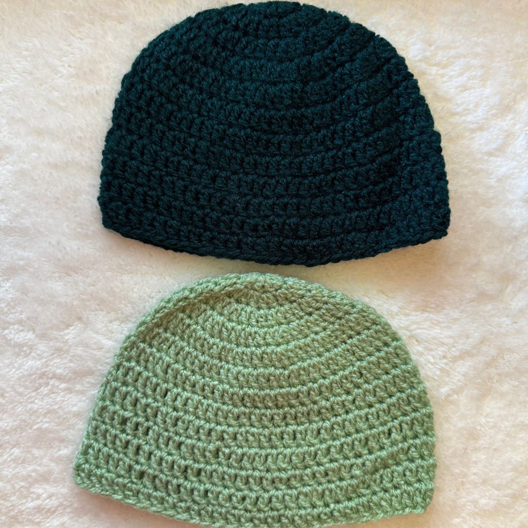 A pair of hand crocheted baby beanie one in dark green and the other in light green placed on a white fur cloth 