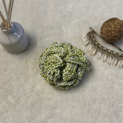 green crochet loofha placed on a soft white fabric with seashells and fragrance sticks in the background