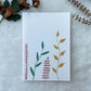 A hand painted greeting card with various leaf stalks painted on a white card placed on a a fur cloth with decorative flowers in the background 