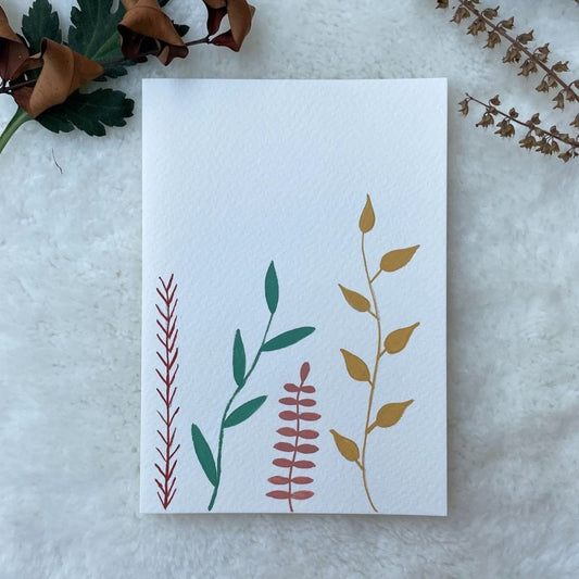 A hand painted greeting card with various leaf stalks painted on a white card placed on a a fur cloth with decorative flowers in the background 