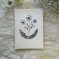 A hand-drawn black and white mandala art and three flowers card placed on a white furry surface, with the card's design visible in the center.