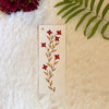 Tiny red flowers wiht orange leaves painted on a bookmark placed on a white fur fabric with fresh flowers and leaves in the background 