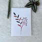 A hand-painted greeting card with a peach and brown gouache illustration of small leaves and grey blossoms on a white background, displayed on a fluffy surface with a small branch of leaves to the side.