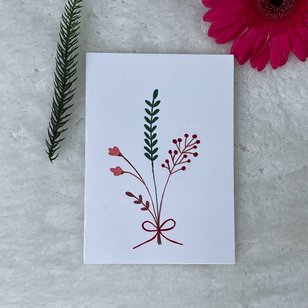 A hand-painted greeting card with a peach minimalist bouquet gouache illustration of small leaves on a white background, displayed on a fluffy surface with a small branch of leaves to the side.