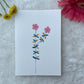 A hand painted greeting card with various flower stalks painted on a white card placed on a a fur cloth with decorative flowers in the background 