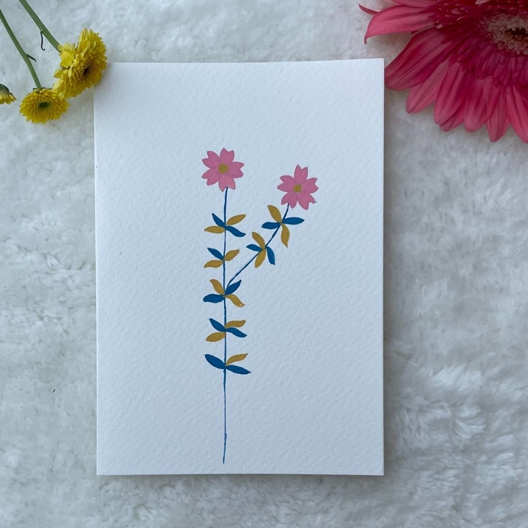 A hand painted greeting card with various flower stalks painted on a white card placed on a a fur cloth with decorative flowers in the background 