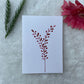 A hand-painted greeting card with a peach and brown gouache illustration of small leaves on a white background, displayed on a fluffy surface with a small branch of leaves to the side.