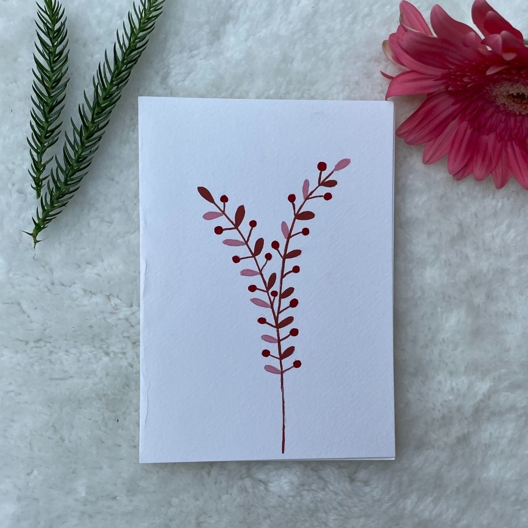 A hand-painted greeting card with a peach and brown gouache illustration of small leaves on a white background, displayed on a fluffy surface with a small branch of leaves to the side.