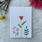 A hand painted greeting card with various flower stalks painted on a white card placed on a a fur cloth with decorative flowers in the background 