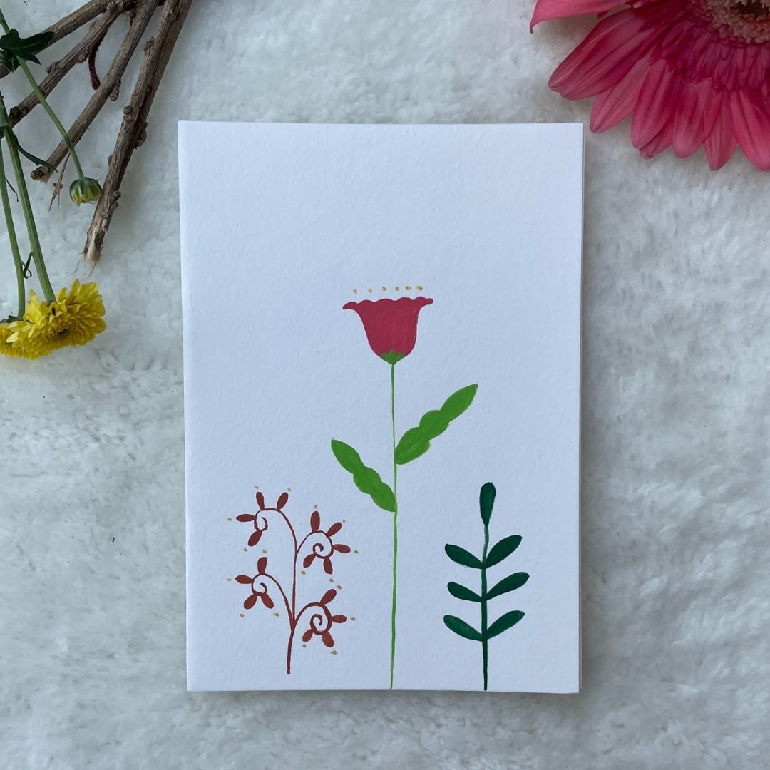 A hand painted greeting card with various flower stalks painted on a white card placed on a a fur cloth with decorative flowers in the background 