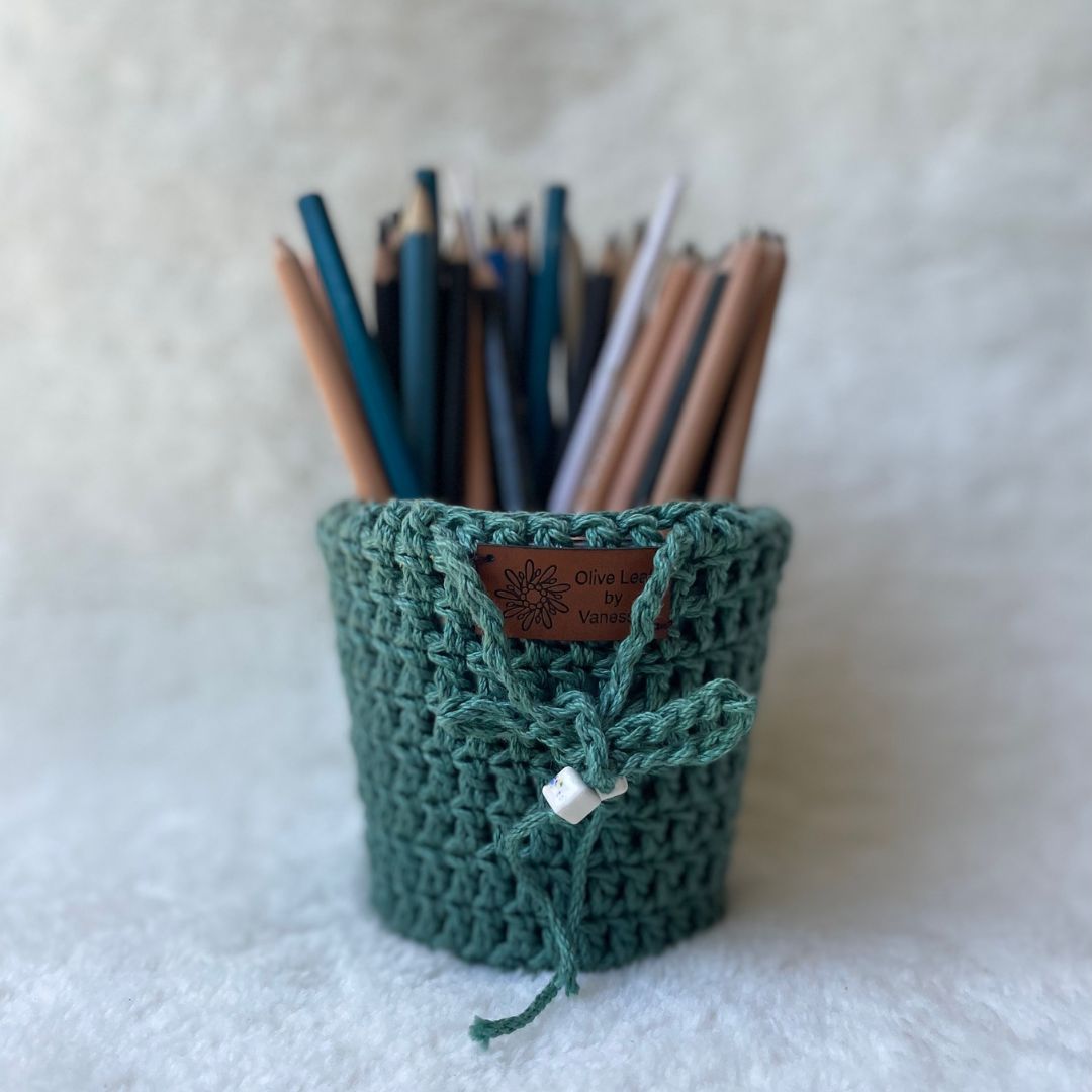 Crochet pot cozy in green holding various pencils placed on a white fur cloth 