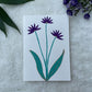 A hand painted greeting card with dark purple flower stalks painted on a white card placed on a a fur cloth with decorative flowers in the background 