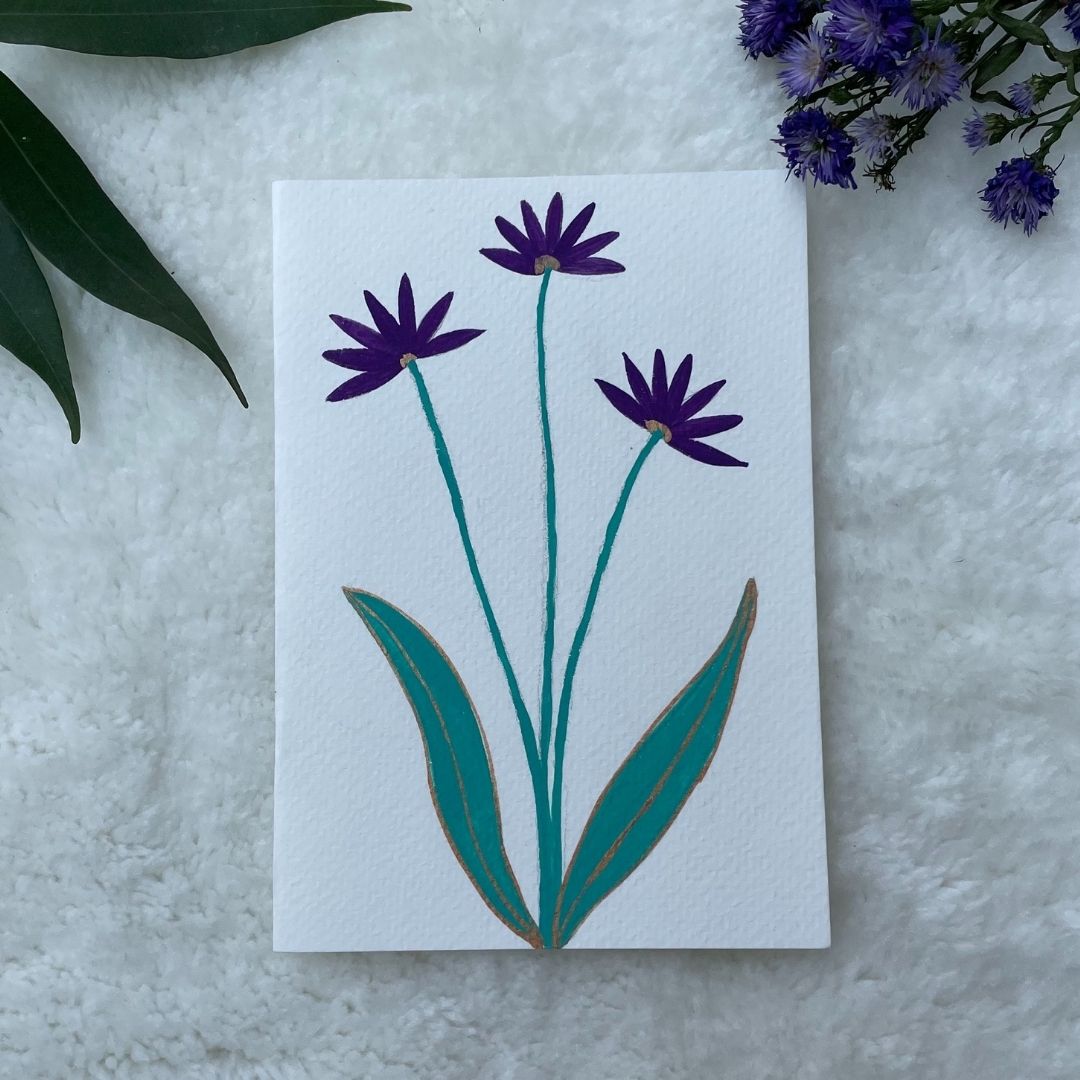 A hand painted greeting card with dark purple flower stalks painted on a white card placed on a a fur cloth with decorative flowers in the background 