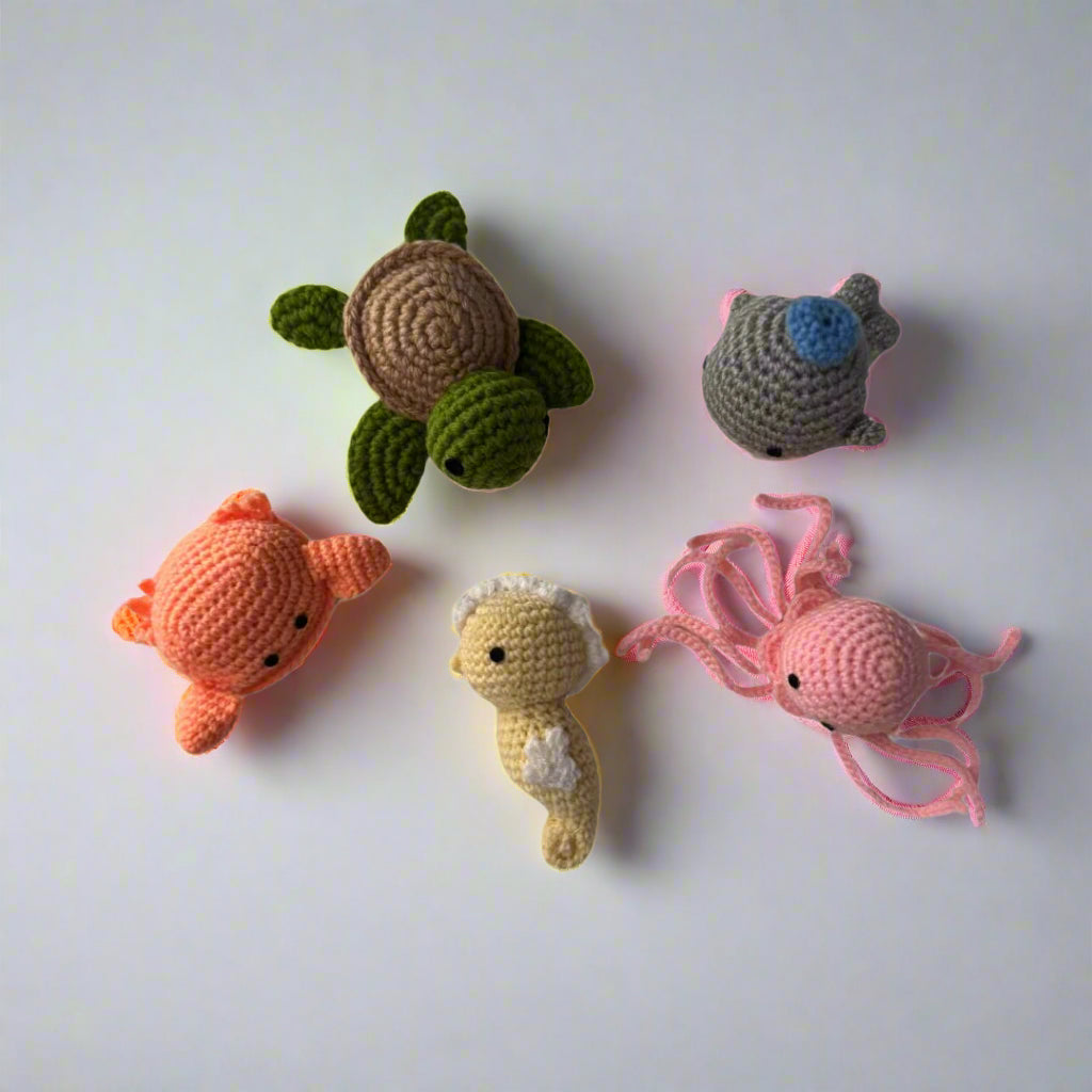 Crochet plushie set of a orange crab, yellow seahorse, grey whale, green turtle and pink octopus placed on a w white table  