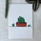 A hand-painted greeting card with illustrations of three plants in pots, placed on a white background with a partial view of a plant to the left and a decorative branch to the top right.