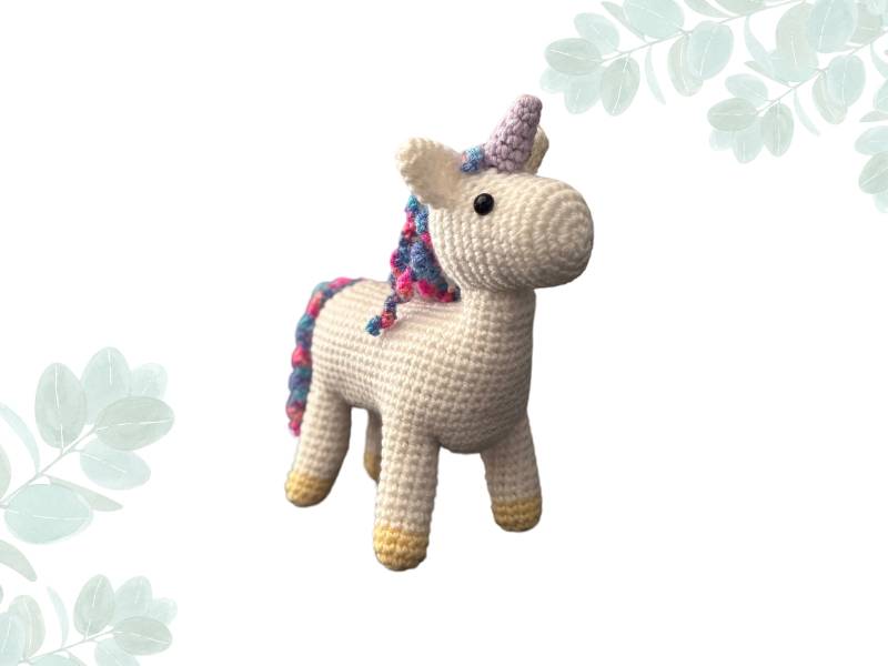 Hand crochet white unicorn with a purple horn and rainbow colour mane and tale, placed diagonally against a white background with leaves