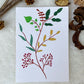 A hand-painted greeting card with a colour leaves and brown blossoms gouache illustration of small leaves on a white background, displayed on a fluffy surface with a small branch of leaves to the side.