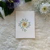 Yellow vinca with green leaves hand painted on a greeting card placed on a soft white cloth with flowers int he background 