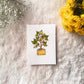 Card with a plant illustration in a yellow pot placed on a textured surface with flowers around