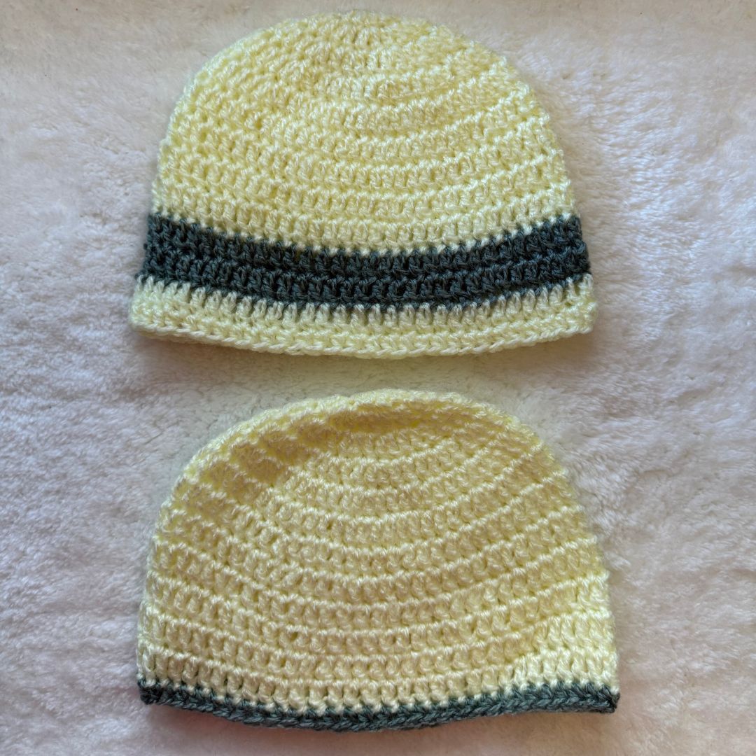 A pair of hand crocheted baby beanies, in yellow with grey stripes placed on a fur cloth 
