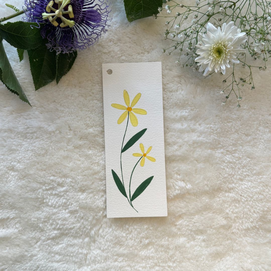 Yellow flowers hand painted on a white bookmark placed on a soft white cloth with flowers in the background 